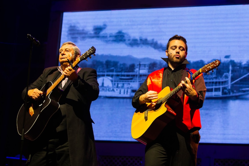 Two men playing guitars on stage, projection of paddle steamer behind