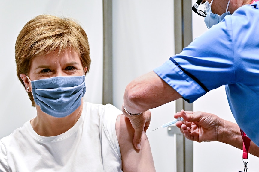 A fair-haired woman sits with her sleeve rolled up as a nurse directs a needle into her arm.