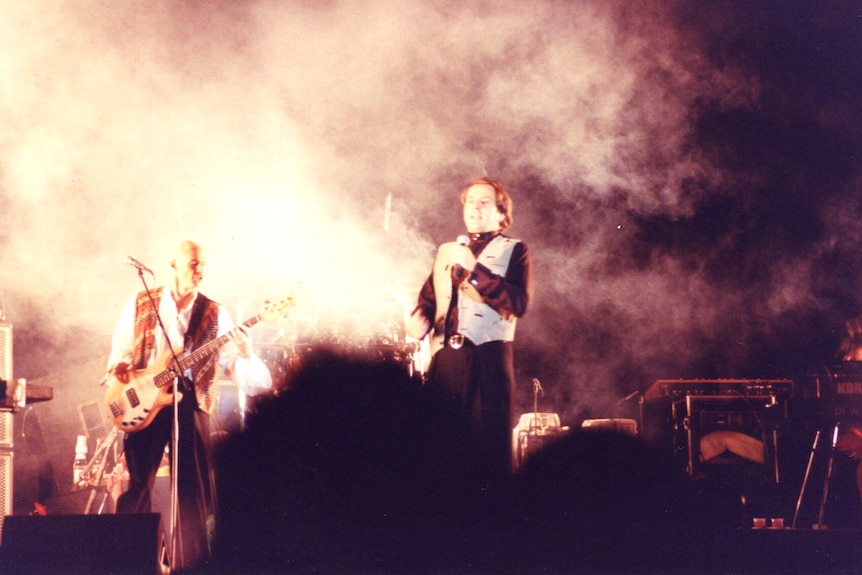 Two men stand on a stage at night time with smoke around them