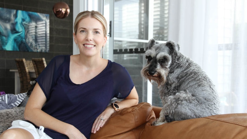 Woman with white shorts and blue shirt sits on couch with dog next to her