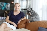 Woman with white shorts and blue shirt sits on couch with dog next to her