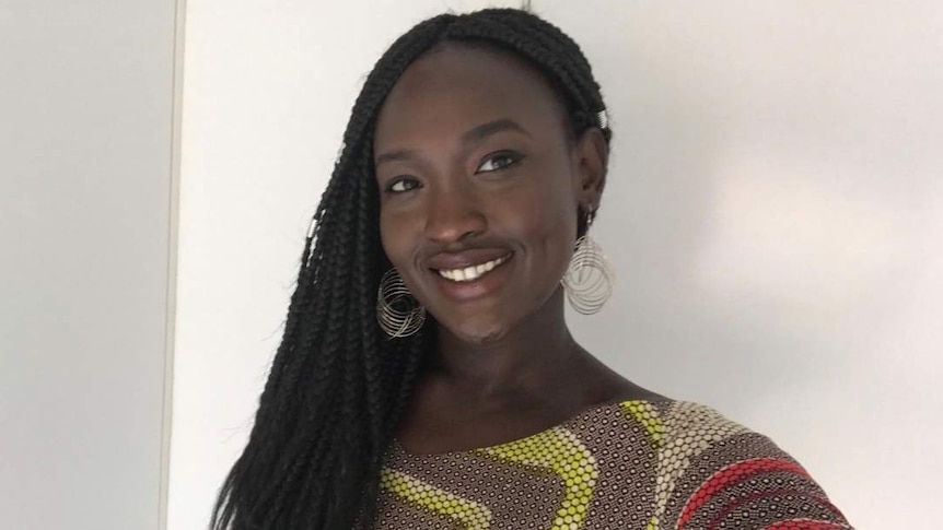 A woman with black skin and plaited hair and a brightly coloured shirt smiles at the camera