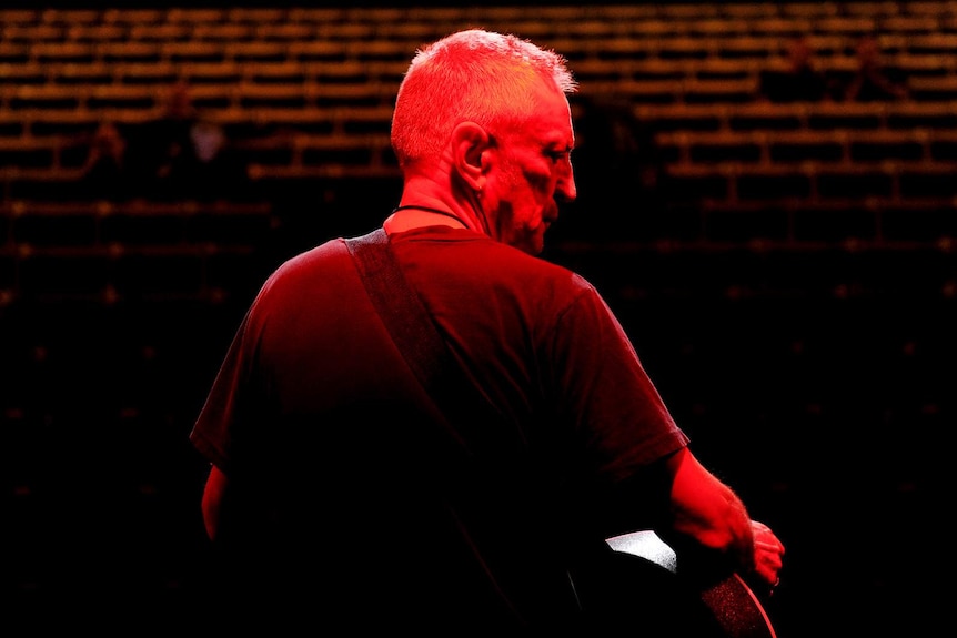 A performer seen from behind, a guitar around his neck, facing a theatre and bathed in red light