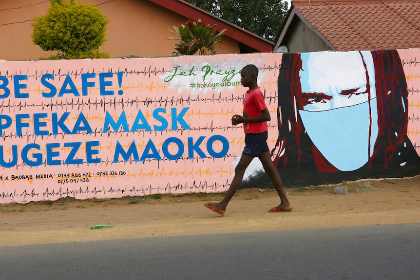 A young boy walks past a wall with pink and blue graffiti urging people to wear face masks with the figure of a man in a mask.
