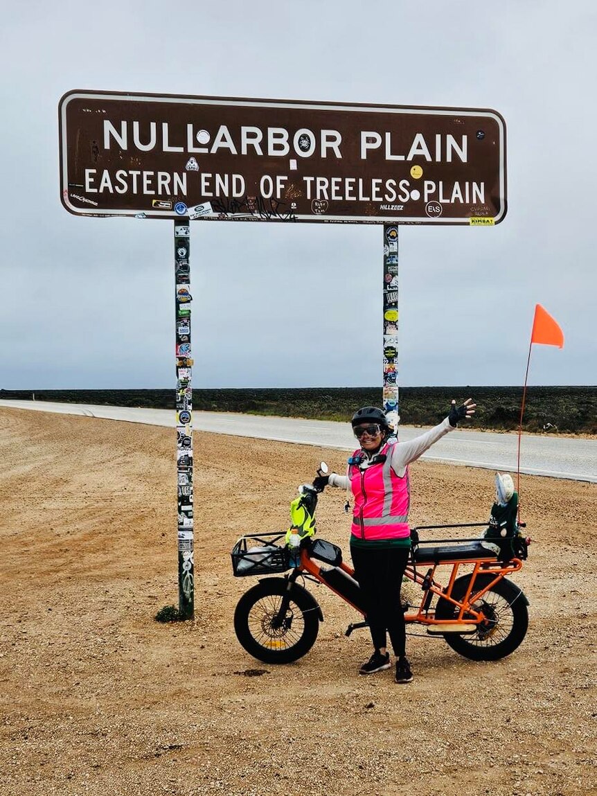 Woman stands by her bike and a sign saying 'Nullarbor Plain, eastern end of treeless plain'