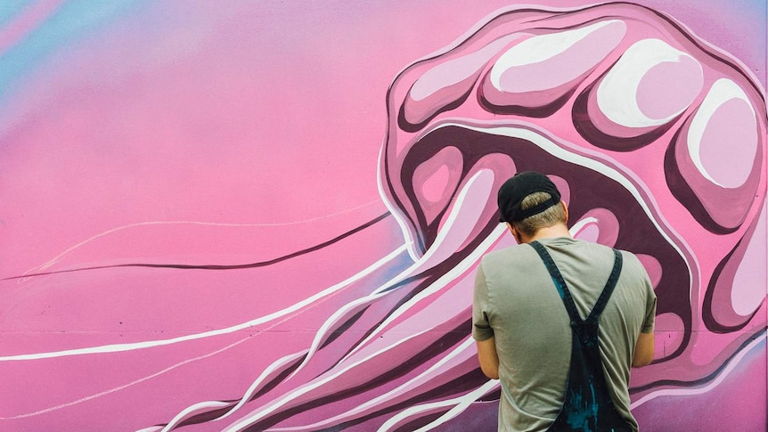 Artwork of a giant pink jellyfish on a wall, with the back of a man wearing blue overalls with paint on them.