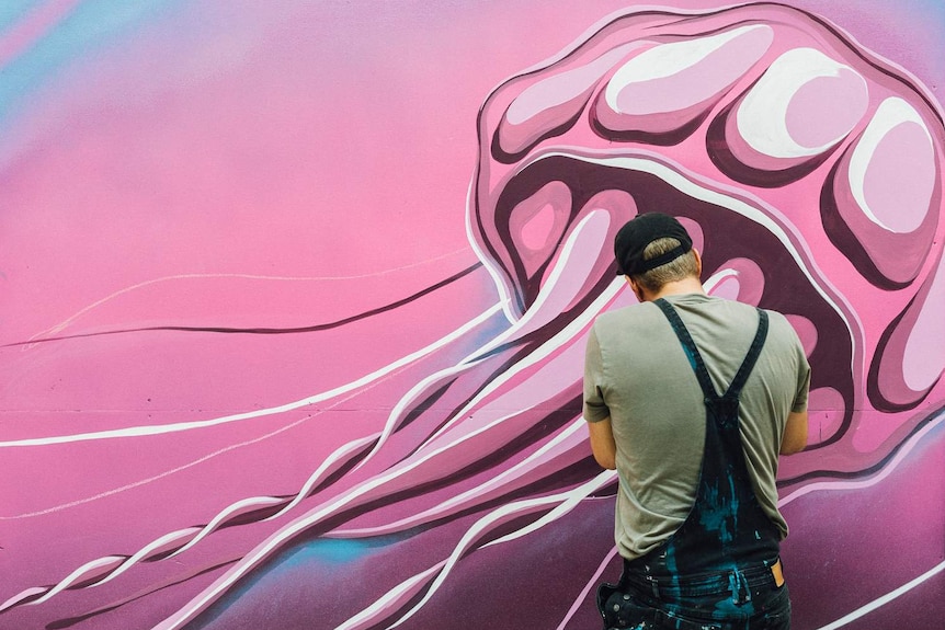 Artwork of a giant pink jellyfish on a wall, with the back of a man wearing blue overalls with paint on them.