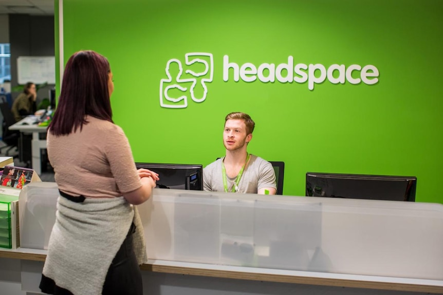 A lady stands at the counter of a headspace centre while a receptionist in a green headspace t-shirt assists.