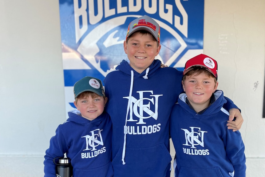 Three young boys in blue hoodies and caps in front of a sign