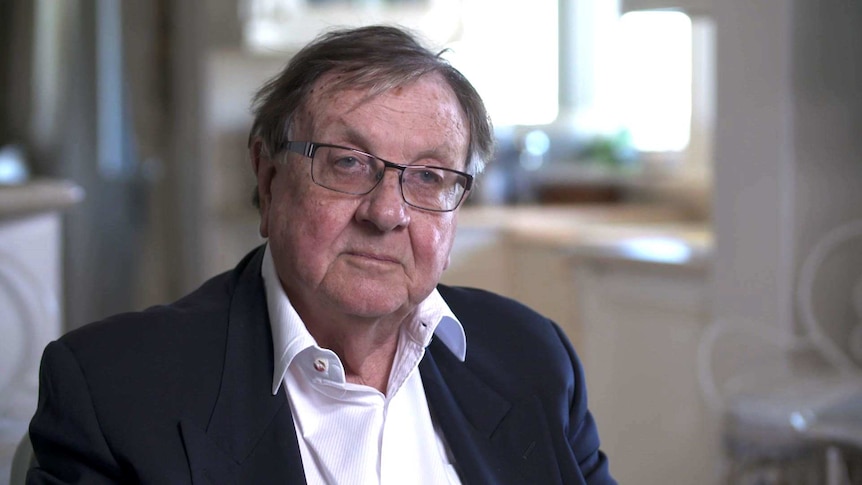 Anthony Whealy wearing glasses, a blue jacket and open collared white shirt sits in his lounge room