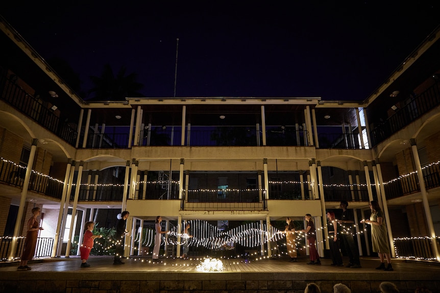 10 people, five on each side of an outdoor stage, hold a string of fairy lights between them. Some swing the lights.