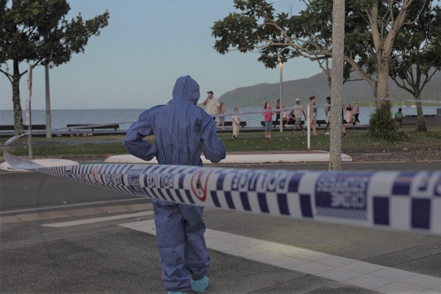 A person in a blue forensics suit walks behind police tape on the Cairns Esplanade