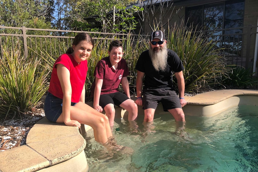 A father and his son and daughter sit on the edge of a backyard pool with their feet in the water.