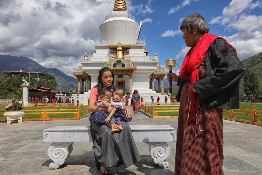 Twin girls in a matching outfits sit on their mother's lap outside a gold-laden temple surrounded by mountainside