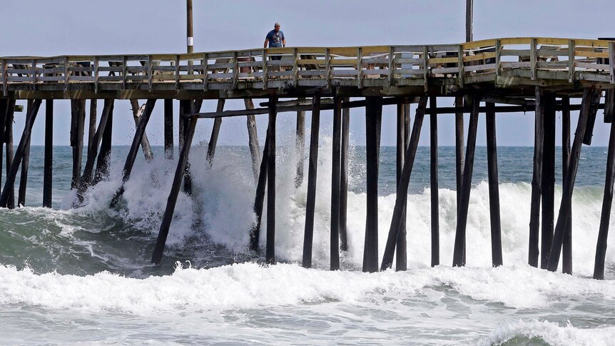 A man walks along a rickety looking wooden wooden pier as big waves crash underneath