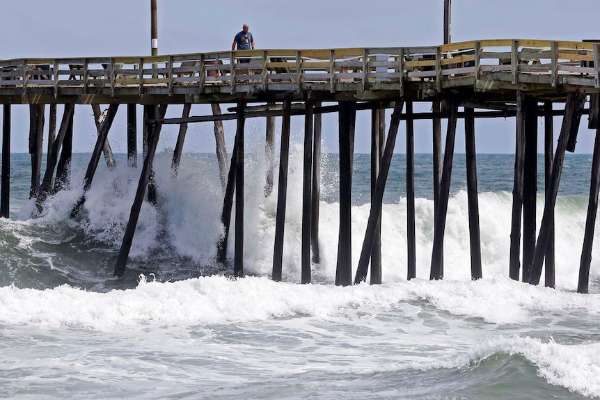 A man walks along a rickety looking wooden wooden pier as big waves crash underneath