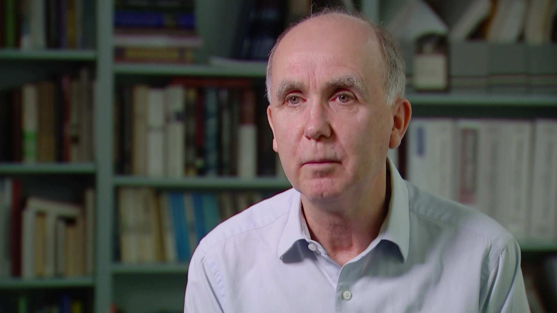 MCU of Jeff Borland wearing a light blue collared shirt, sitting in front of shelves of books