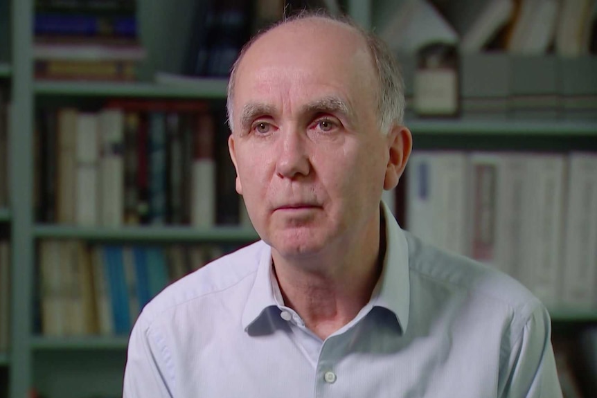 MCU of Jeff Borland wearing a light blue collared shirt, sitting in front of shelves of books