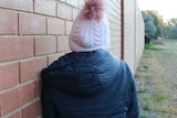 A young person wearing a beanie and hooded jacket stands against a brick wall.
