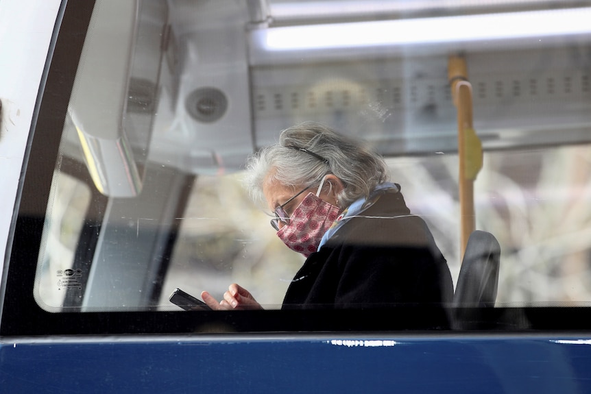 An older woman is photographed in a pink mask through a bus window. 