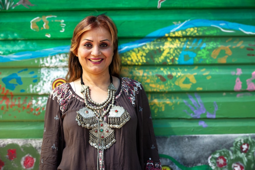 A woman stands in front of a green wall, smiling. She is wearing a traditional Afghan necklace