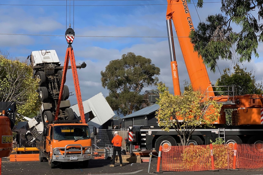 Two orange cranes begin to winch up a toppled crane as people in orange vests and hard hats watch on.