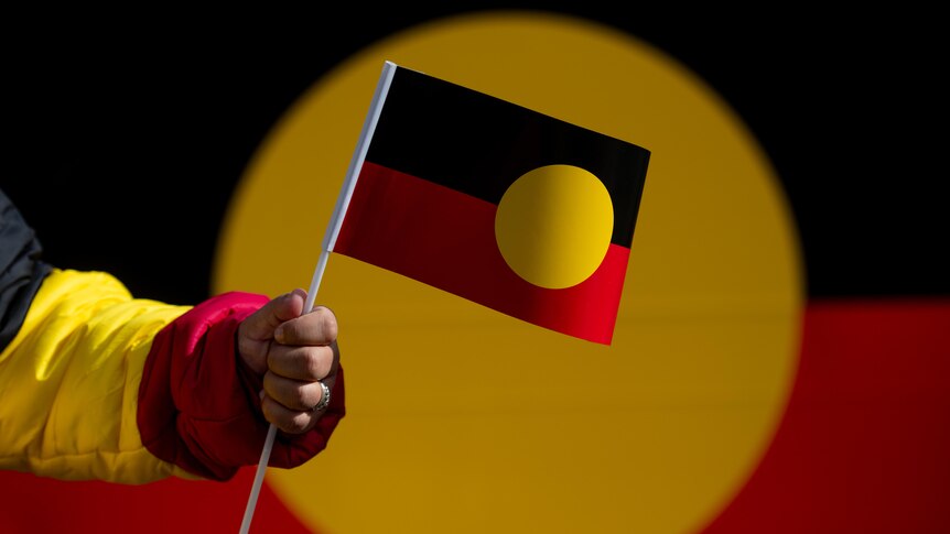 A hand holds an Aboriginal flag in front of a large banner