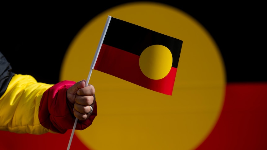 Photo of a person holding up an Aboriginal flag. Behind it, out of foucs, is an even larger Aboriginal flag.