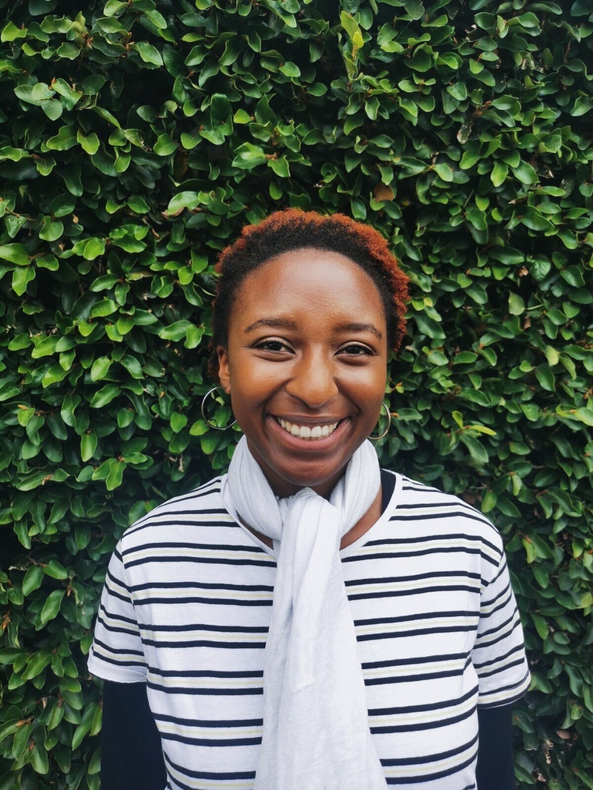 Thandi smiles as she stands in front of a green hedge wearing a striped top, in a story about natural hair movement.