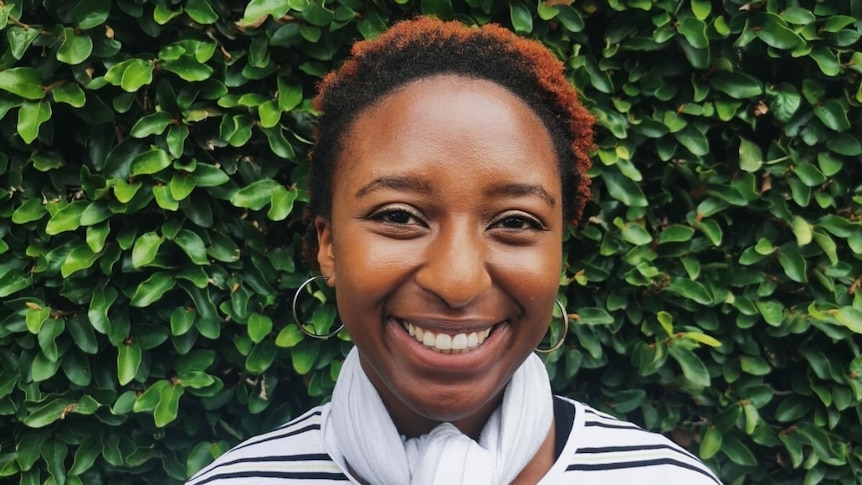 Thandi smiles as she stands in front of a green hedge wearing a striped top, in a story about natural hair movement.