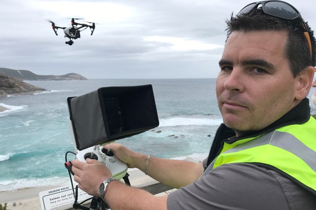 A man turns and looks at the camera with drone controls and a drone in front of him while standing on the coast.