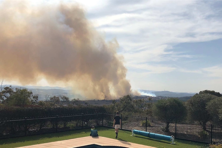A plume of smoke billows up from the fire, a child watching from beside a pool in the foreground.