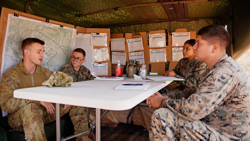 Four US Marines in army camouflage seated around a tabled in a tent, with documents pinned to the walls behind them