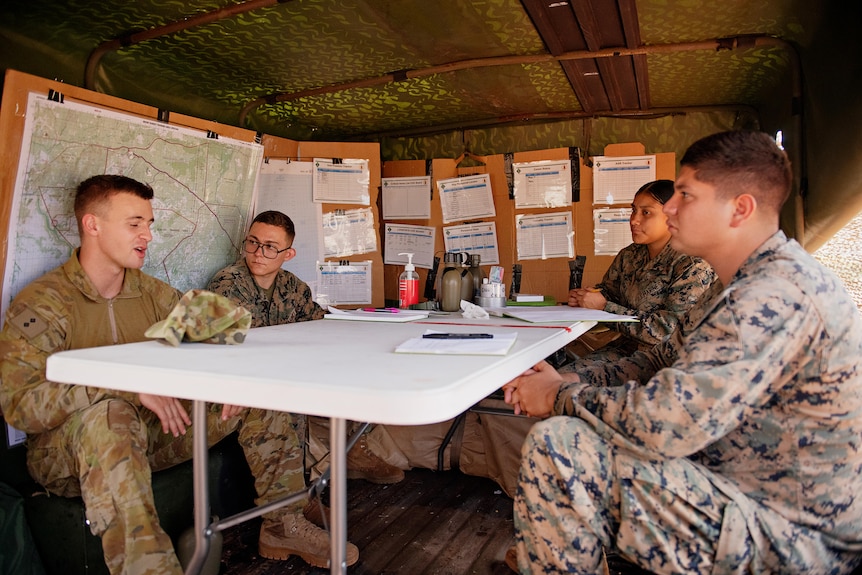 Four US Marines in army camouflage seated around a chalkboard in a tent, with documents pinned to the walls behind them
