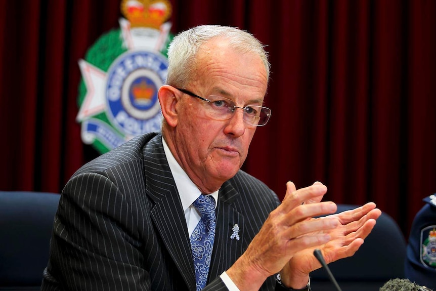 Bob Atkinson, wearing a suit with the Queensland police logo behind him on a wall, speaks at a press conference in 2011.