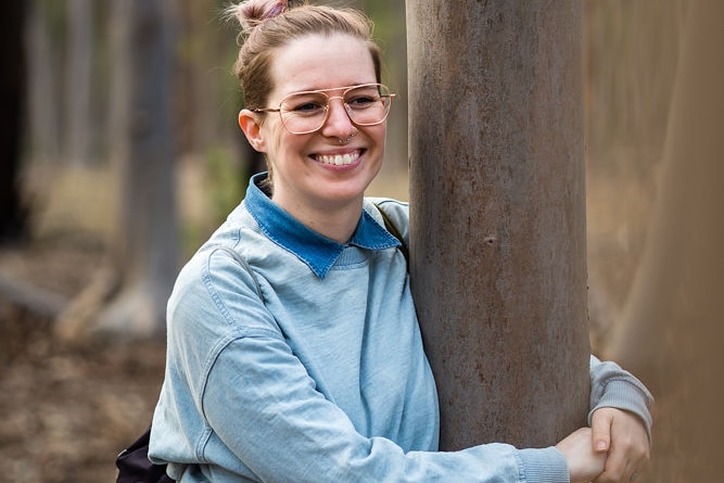 A woman with wide smile and glasses wraps her arm around a tree.
