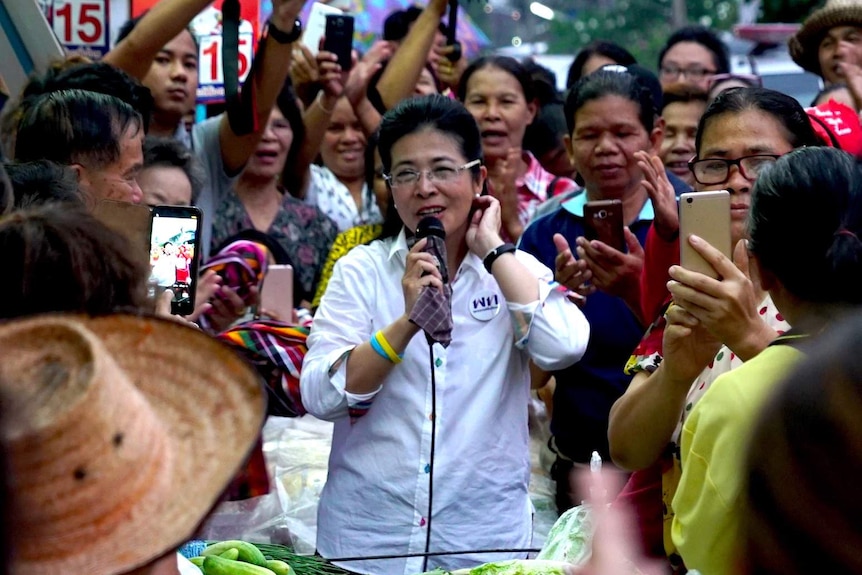 A woman holds a microphone surrounded by people at a market in Thailand