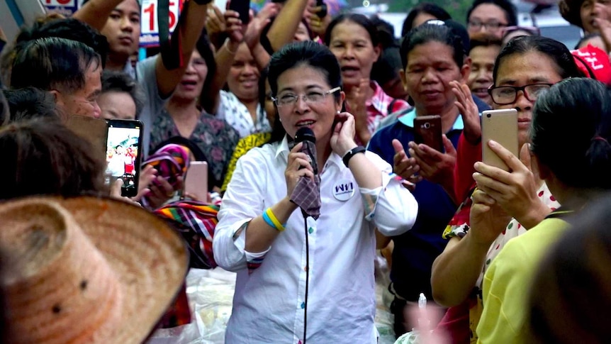 A woman holds a microphone surrounded by people at a market in Thailand