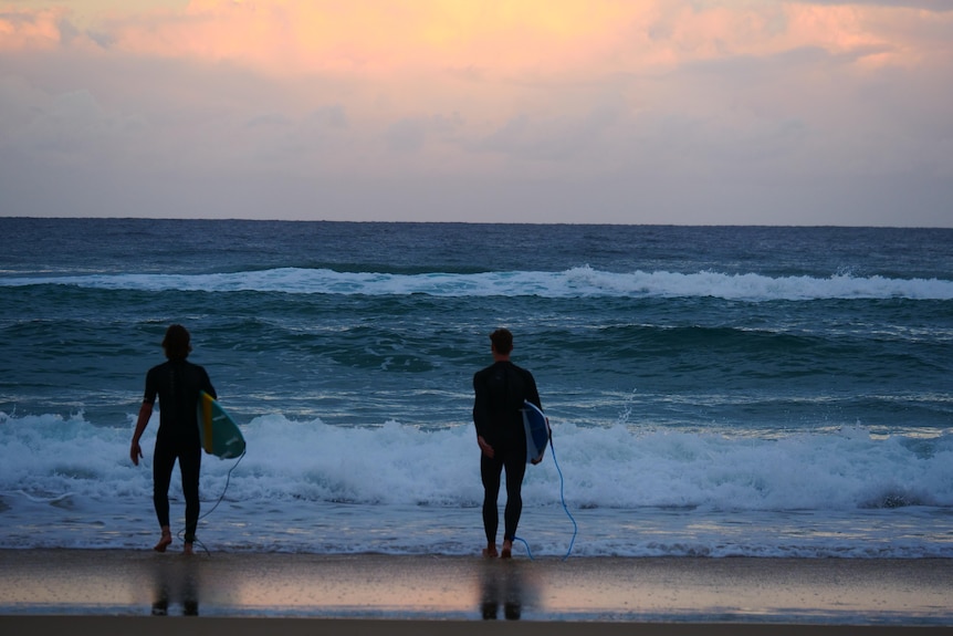 A pair of surfers stand on the tideline in low light.