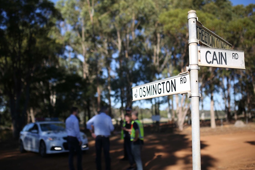 A street sign showing the names of Osmington Road and Cain Road on a rural dirt road with police officers and a car behind.