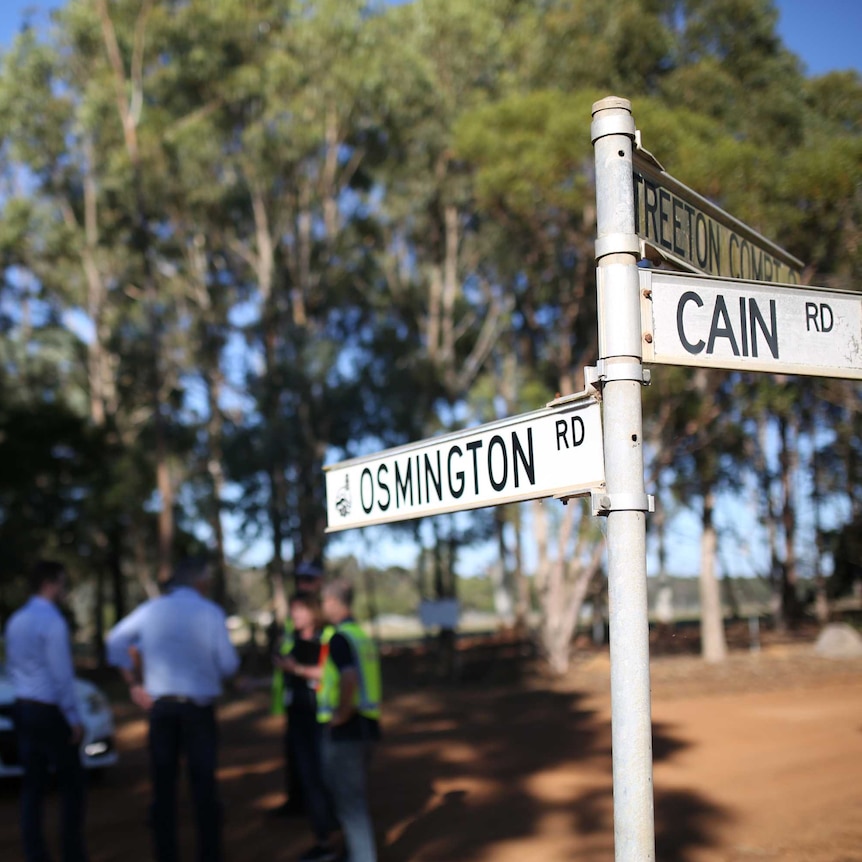 A street sign showing the names of Osmington Road and Cain Road on a rural dirt road with police officers and a car behind.