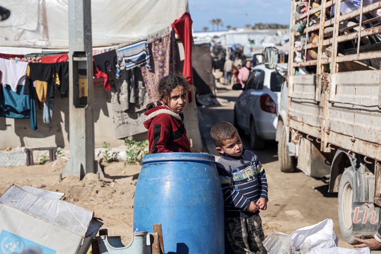 Two young children standing near a barrel, looking at the camera with neutral expressions on their faces.