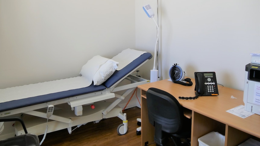 A medical examination bed is next to a desk with a phone, printer.