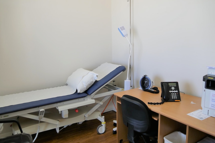 A medical examination bed is next to a desk with a phone, printer.