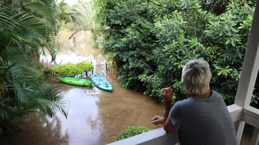 A woman in a grey shirt leans on her balcony, looking at murky brown floodwaters that have nearly risen to the top of her fence.