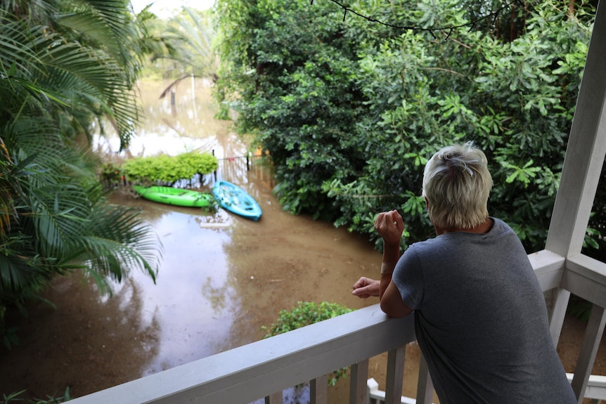 A woman in a grey shirt leans on her balcony, looking at murky brown floodwaters that have nearly risen to the top of her fence.