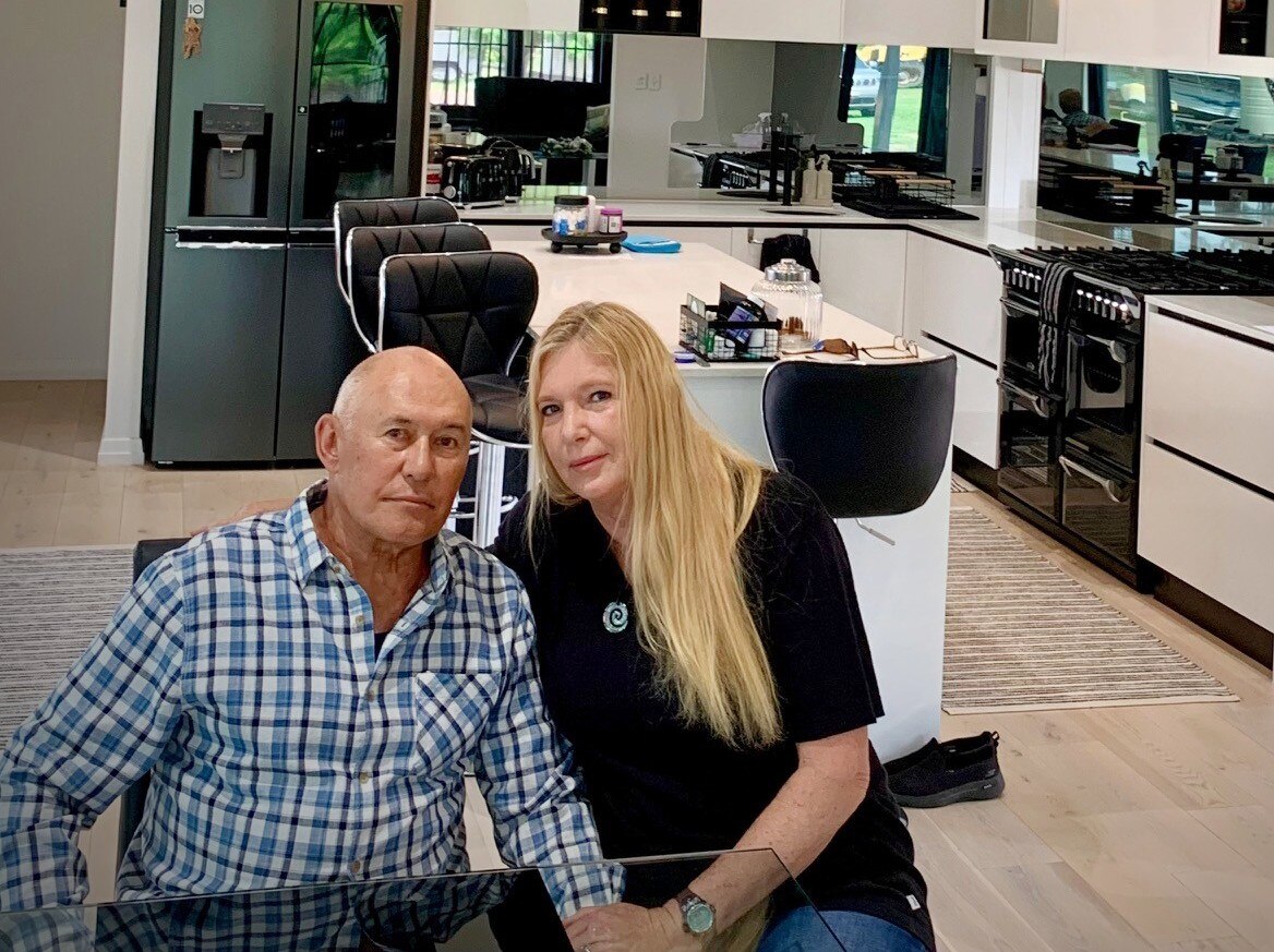 A man in a checked shirt sitting with a blonde woman in a black polo at a table in front of a kitchen setting