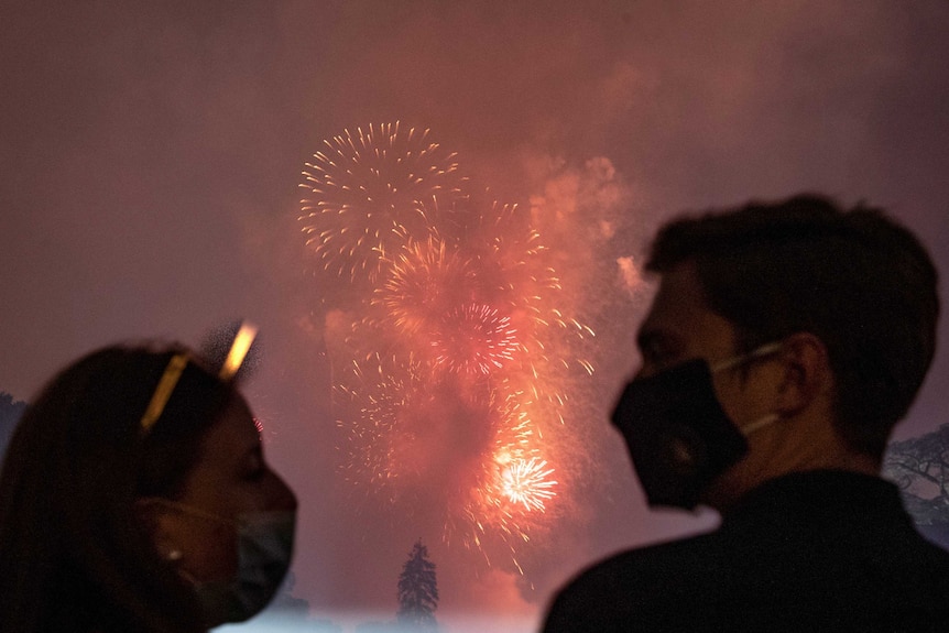 Two people with face masks in foreground watch fireworks display with trees in background.