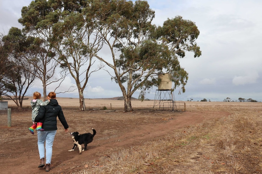 Farm scene with trees and paddock and woman with daughter on hip walking away from camera with dog