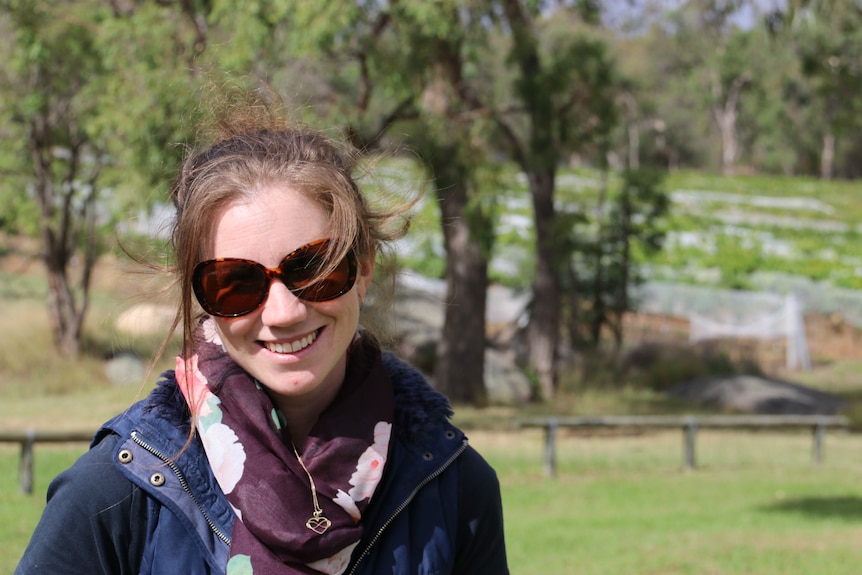 A woman with brown hair in a navy raincoat smiles at the camera with trees in the background